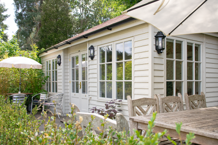 Hipped Garden Room with internal partitions by Crane Garden Buildings Traditional garden room with painted timber cladding, French doors and large windows, surrounded by a furnished garden terrace and planting.